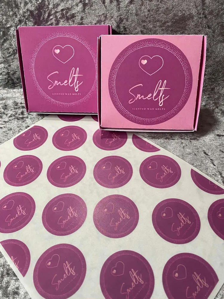 Pink logo-printed cardboard wax melt box beside a sheet of round logo prints